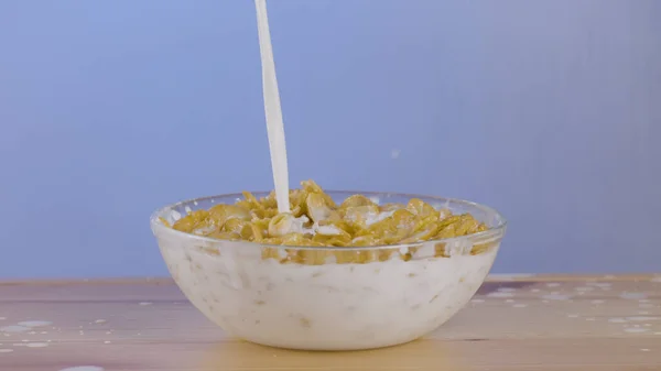 Pouring milk over a transparent bowl full of cornflakes with drops of milk falling on a table. Action. Tasty breakfast with cereal. — Stock Photo, Image