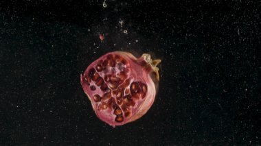 Siyah zemin üzerinde beyaz küçük parçacıklarla suya düşen narı kapat. Başla. Suyun altında hareket eden olgun bir meyve, vitamin konsepti ve sağlıklı beslenme. 