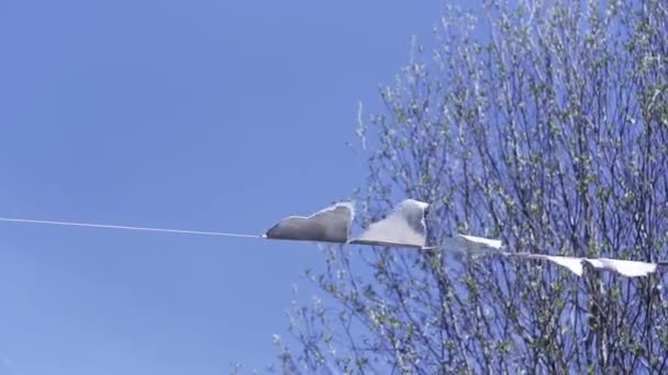 Onderaanzicht van driehoekige witte vlaggen zwaaiend in de wind. Een knip. Mooi buitenshuis vakantie decoratie op blauwe lucht en boom takken achtergrond. — Stockvideo