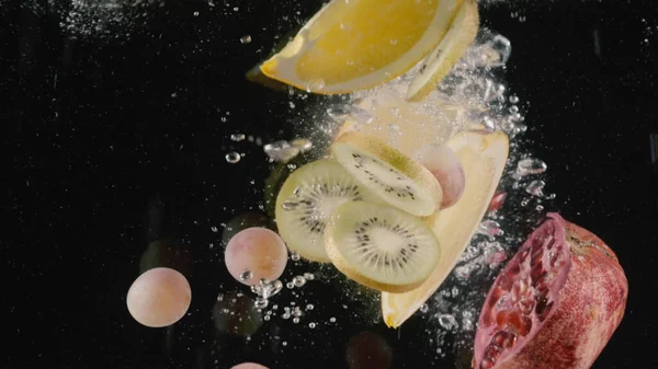 Крупным планом лимона, ломтиков киви, винограда и половины граната, брошенного в воду изолированы на черном фоне. Начали. Плоды и ягоды в прозрачной воде с пузырьками воздуха, концепция — стоковое фото