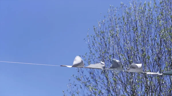 Bottom view of triangular white flags swaying in the wind. Clip. Beautiful outdoors holiday decoration on blue sky and tree branches background.