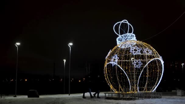 Nachtansicht von zwei Silhouetten, die um eine wunderbare riesige Spielzeugkugelfigur herumlaufen, die mit leuchtenden LED-Lichtern dekoriert ist. Konzept. Nachtwinterpark mit Laternen und Weihnachtsdekoration. — Stockvideo