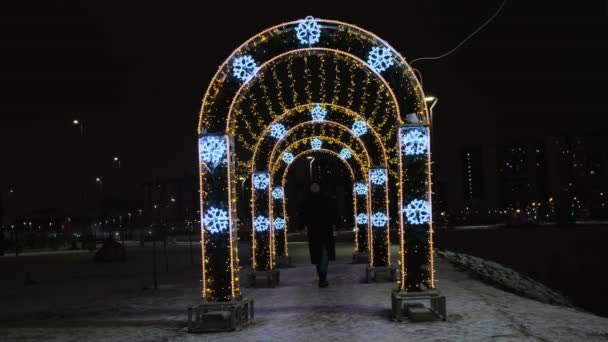 Capodanno decorazione luminosa di Natale nel parco della città su sfondo cielo nero. Concetto. Un uomo che attraversa il tunnel luminoso con ghirlande dorate e fiocchi di neve blu. — Video Stock