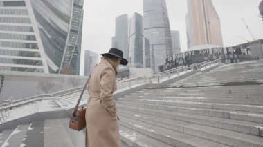 Kahverengi ceketli ve siyah şapkalı güzel iş kadını modern şehir bölgesinde gökdelenleri olan üst kata çıkıyor. Başla. Güzel cam cephe binalarının yanında yürüyen bir kadının yan görüntüsü.. 