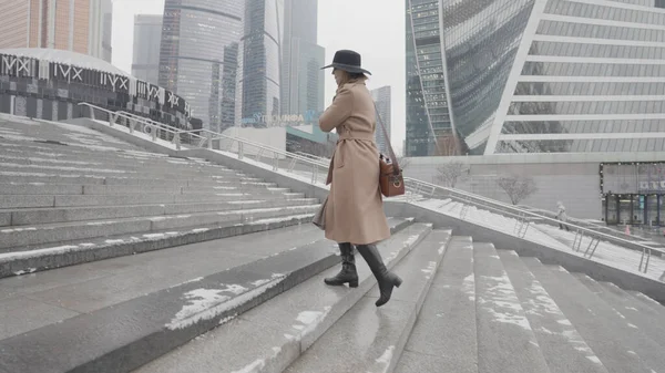 Hermosa mujer de negocios con un abrigo marrón y sombrero negro que va arriba en el moderno distrito de la ciudad con rascacielos. Acción. Vista lateral de una mujer caminando cerca de hermosos edificios de fachada de vidrio. — Foto de Stock