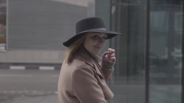 Oldalnézetben egy elegáns, boldog nő kabátban és kalapban sétál az utcán. Felvétel. Nevető nő levesz egy kalapot az erős szél miatt.. — Stock videók
