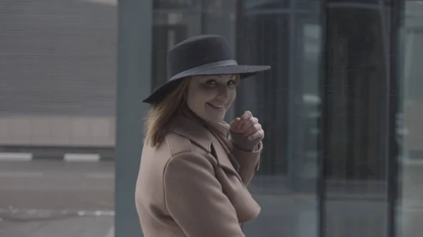 Seitenansicht einer eleganten, glücklichen Frau in Mantel und Hut, die auf der Straße spaziert. Handeln. Lachende Frau nimmt wegen starken Windes ihren Hut. — Stockfoto