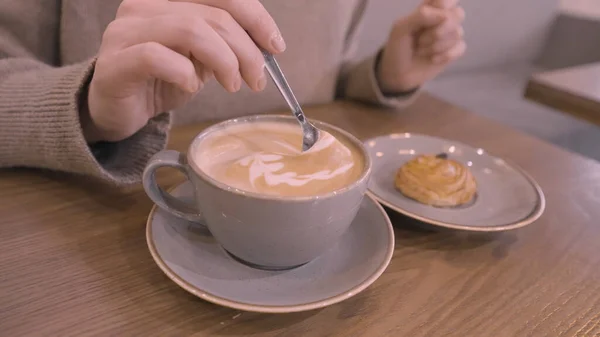 Mexendo café com leite com uma colher e um biscoito deitado em um prato. Acção. Close up de mão mulher mexendo espuma de seu café enquanto sentado no restaurante. — Fotografia de Stock