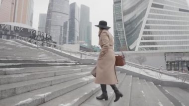 Kahverengi ceketli ve siyah şapkalı güzel iş kadını modern şehir bölgesinde gökdelenleri olan üst kata çıkıyor. Başla. Güzel cam cephe binalarının yanında yürüyen bir kadının yan görüntüsü.. 