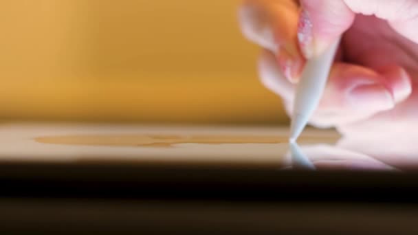 Nahaufnahme einer Mädchenhand mit einem weißen Stift zum Zeichnen auf einem digitalen Tablet-Gerät. Konzept. Frau zeichnet ein Bild auf einem Pad auf verschwommenem gelben Hintergrund. — Stockvideo