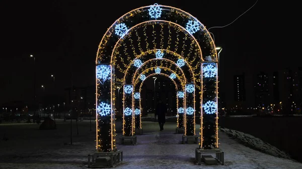 New Year Christmas luminous decoration in the city park on black sky background. Concept. A man passing through the luminous tunnel with golden garlands and blue snowflakes. — Stock Photo, Image