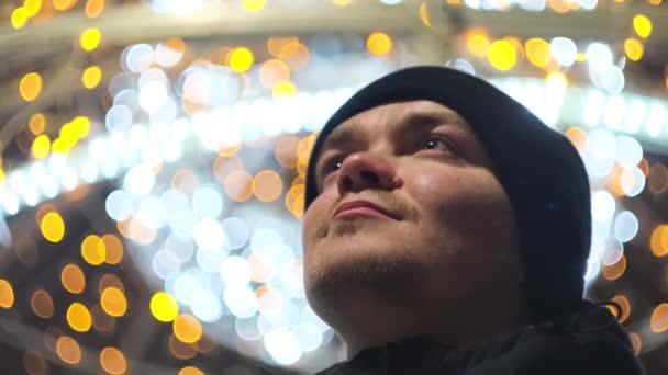 Blick von unten auf einen jungen, glücklichen Mann, der im abendlichen kalten Wetter strahlende Weihnachtsgirlanden im Freien betrachtet. Konzept. Lächelnder Mann bewundert festliche Stadtdekorationen vor verschwommenem Glanz — Stockvideo