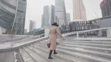 Soğuk bir havada, uzun kahverengi paltolu ve siyah şapkalı bir kadın şehir caddesinde yürüyor. Başla. Genç bir kadın yüksek gökdelenlerin önünde merdivenlerden çıkıyor..