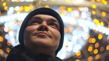 Genç ve mutlu bir adamın akşam soğuğunda dışarıda parlayan Noel çelenklerini izlemesi. Kavram. Gülümseyen adam bulanık arka plandaki şenlikli şehir dekorasyonlarına hayran.
