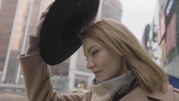 Egy fiatal szőke nő portréja meleg barna kabátban és sálban, fekete kalapban. Felvétel. Nő áll a szabadban a város utcájában előtt felhőkarcolók. — Stock videók