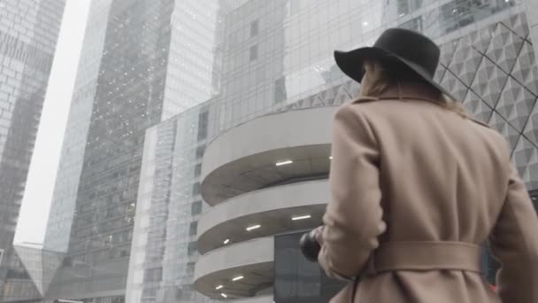 Attraente giovane donna bionda con una macchina fotografica tra le mani nella strada della città. Azione. Elegante femmina in cappotto marrone e cappello nero sorridente e in posa in una giornata invernale sullo sfondo di edifici moderni — Video Stock