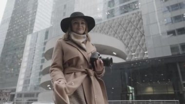 Şehir caddesinde elinde kamera olan çekici sarışın bir kadın. Başla. Kahverengi ceketli, siyah şapkalı, kışın gülümseyen ve poz veren zarif kadın modern binaların arka planında.