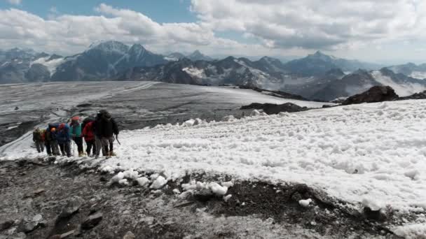 Jóvenes escaladores en chaqueta abrigada, cascos y gafas especiales subiendo en una ladera de montaña uno por uno en una fila. Clip. Personas que se levantan en la pendiente cubierta de nieve en el fondo de muchas montañas y azul — Vídeo de stock