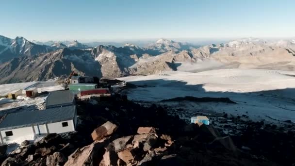 雪の岩やハイカーのための避難所と自然景観。クリップ。山頂からの眺めは、山脈を背景にした登山者のための小さなキャンプ場と青空の背景. — ストック動画