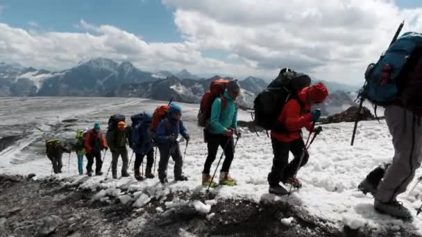 Sıcak ceketli genç tırmanışçılar, miğferler ve özel gözlükler dağ yamacında sırayla ilerliyorlar. Şarjör. Karda yükselen insanlar arka planda birçok dağ ve mavi yamaçta uzanır. — Stok video