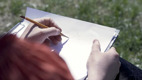 Close up de artista mulher desenhando algo com um lápis em um caderno de esboços ao ar livre. Clipe. Sobre a vista do ombro de desenhar um quadro enquanto sentado em um prado verde. — Fotografia de Stock