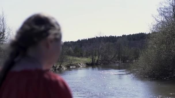 Bakre bild av en medelålders naturlig kvinna i en röd skjorta med lång fläta framför strömmande flod och sommarskog. Klipp. Begreppet enhet med naturen. — Stockvideo