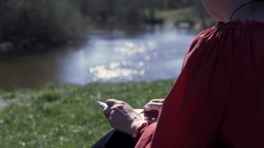 Nehir ve yaz ağaçlarının önündeki yeşil tepede oturan bir kadın. Şarjör. Beyaz bir kağıt üzerine çizim yapmak için kalem kullanan kadın, doğal bir ilham kaynağı.. 