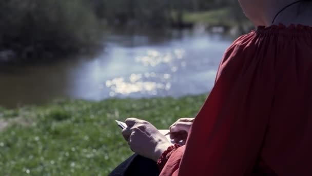 Mujer sentada en una colina verde frente a un río y árboles de verano. Clip. Mujer usando lápiz para dibujar un boceto en una hoja blanca de papel, inspiración natural. — Vídeos de Stock