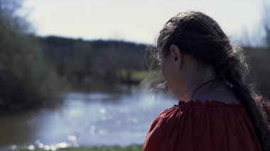 Orta yaşlı, kırmızı tişörtlü, nehir ve yaz ormanlarının önünde uzun örgülü doğal bir kadının arka görüntüsü. Şarjör. Doğayla birlik kavramı.