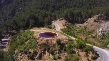 Dağ yamacında bulunan yuvarlak, koyu kırmızı bir gölün havadan görünüşü. Şarjör. Ormanlık yeşil tepeleri ve dar beton yolu olan güzel yaz manzarası..