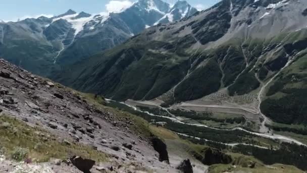 Increíble paisaje con montañas gigantes con pendientes verdes y picos nevados. Clip. Sendero pedregoso a lo largo de rocas cubiertas por vegetación verde sobre fondo azul nublado del cielo. — Vídeos de Stock