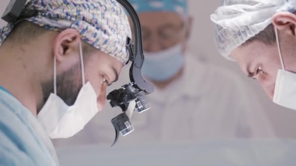 Docteur et un assistant dans la salle d'opération de la clinique de chirurgie vasculaire veineuse chirurgicale. L'action. Vue latérale d'un chirurgien avec une loupe binoculaire. — Video