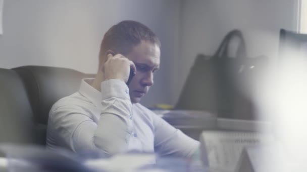 Podnikatel pracuje ve své kanceláři před počítačovou obrazovkou. Akce. Boční pohled na muže v košili, jak mluví na mobilním telefonu o práci. — Stock video