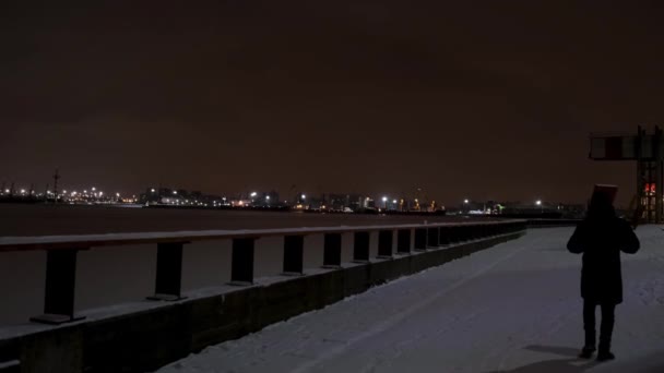 Rückansicht eines Mannes, der nachts auf einer schneebedeckten Straße die Böschung entlang geht. Konzept. Männchen läuft allein bei kaltem Wetter mit der Stadtbeleuchtung im Hintergrund. — Stockvideo
