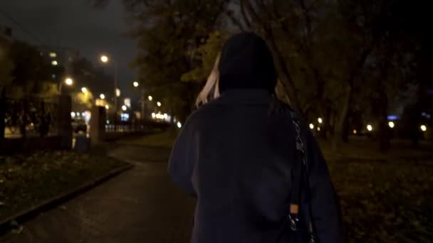 Achteraanzicht van een mooi meisje dat 's nachts door het park gaat, ze is bang en begint te rennen. Actie. Blond tiener meisje op zoek bang in het stadspark laat in de avond. — Stockvideo