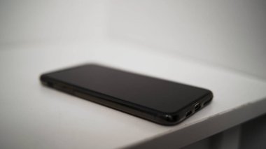 Erkek eli siyah cep telefonunu beyaz arka planda izole bir masaya koyar. Başla. Erkek eli kapalı, akıllı telefonu masaya koyuyor..