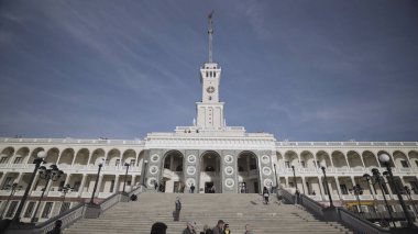 Mavi bulutlu arka planda beyaz bir tren istasyonu ana binasının alt manzarası. Başla. Güzel bir binanın yanında yürüyen insanlar, mimari konsepti..