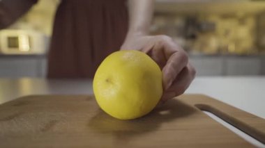 Evin mutfağındaki ahşap bir tahtanın üzerinde sarı limonu çelik bıçakla kesen kadın ellerine yakın çekim. Başla. Limonata hazırlama süreci.