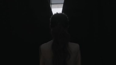 Rear view of a young woman with long curly hair opening curtains on window in the morning at home. Action. Silhouette of a girl in the dark room opening curtains.