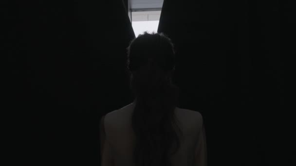 Rear view of a young woman with long curly hair opening curtains on window in the morning at home. Action. Silhouette of a girl in the dark room opening curtains. — Stockvideo