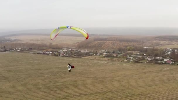 Mannen med en mantel som flyger på en paraglider som en Stålman över ett gult höstfält. Börja. Flygande fallskärmshoppare nära byn en dimmig dag. — Stockvideo