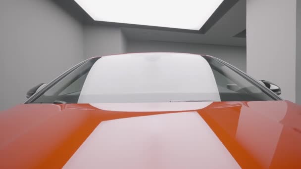 Primer plano de la vista frontal de un coche deportivo de lujo rojo estacionado en el garaje. Acción. Parabrisas y capó del coche bajo el reflector brillante. — Vídeo de stock