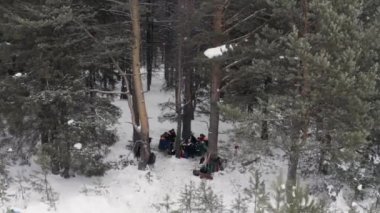 Erkek avcıların kış ormanlarında ateşin etrafında oturduğunu ve dinlendiğini gösteren hava manzarası. Şarjör. Soğuk bir kış gününde ısınan bir grup insan.