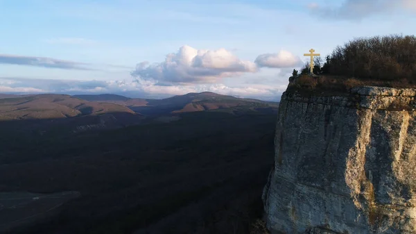 Золотой крест на вершине горы с крутым склоном, концепция религии. Выстрел. Вид с воздуха на скалу и красивую холмистую долину на заднем плане. — стоковое фото