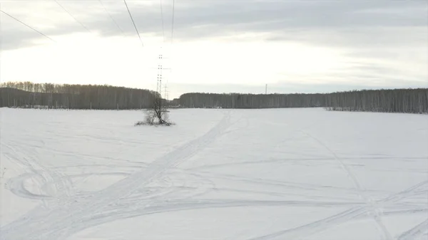 Aerial view of a high voltage transmission tower at snow covered field with trees on a winter day. Clip. White field with traces of snowmobiles on forest background. — Stock Photo, Image