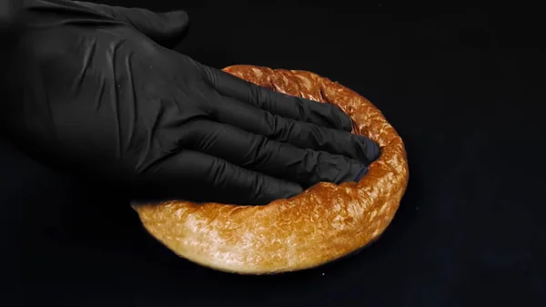 Close up of hand in black cooking glove touching fresh burger round bun with gergelim seeds. Imagens de stock. Mão pressionando o pão para mostrar sua frescura no fundo preto. — Fotografia de Stock