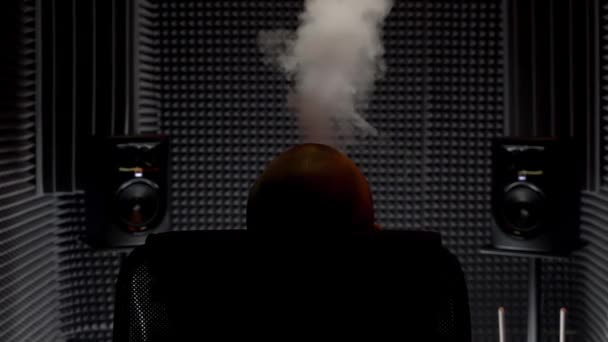 Visão traseira de um homem sentado em uma poltrona exalando fumaça em um estúdio de gravação de música. HDR. Músico masculino relaxante, fumante ou vaping em um estúdio com paredes de isolamento acústico. — Vídeo de Stock