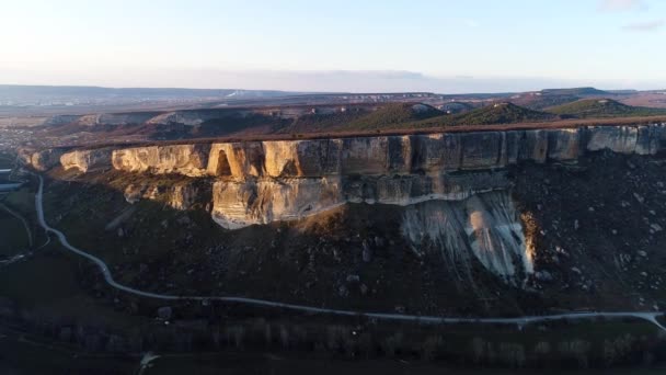 Widok z lotu ptaka gigantycznego pięknego klifu w świetle zachodu słońca. Postrzelony. Naturalny krajobraz zapierającej dech w piersiach skały z płaskim wierzchołkiem otoczony zielonymi wzgórzami i wąską drogą. — Wideo stockowe