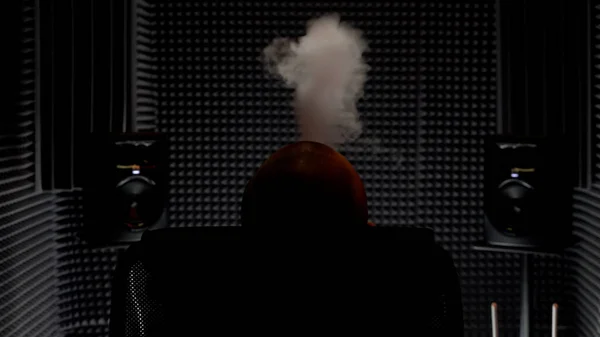 Achteraanzicht van een man die in een fauteuil rook uitademt in een muziekopnamestudio. HDR. Mannelijke muzikant ontspannen, roken of dampen in een studio met geluidsisolatie muren. — Stockfoto