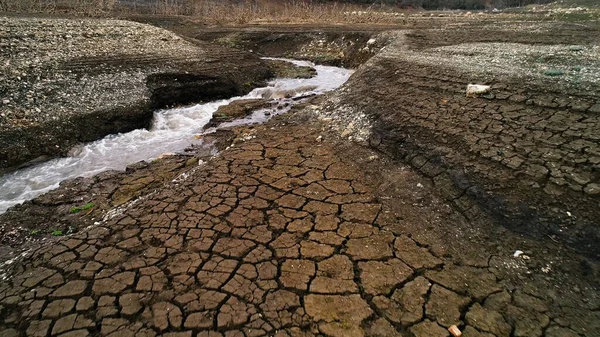 Landscape of dry earth ground and river with water streaming. Shot. Aerial view of a field with cracks and a narrow river, concept of ecology and global warming. — Stock Photo, Image
